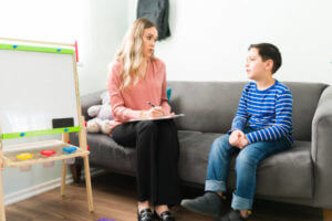 Caucasian female therapist in her 30s talking about elementary school with a little boy. Happy boy sitting on the sofa and having a chat with a psychologist at her office